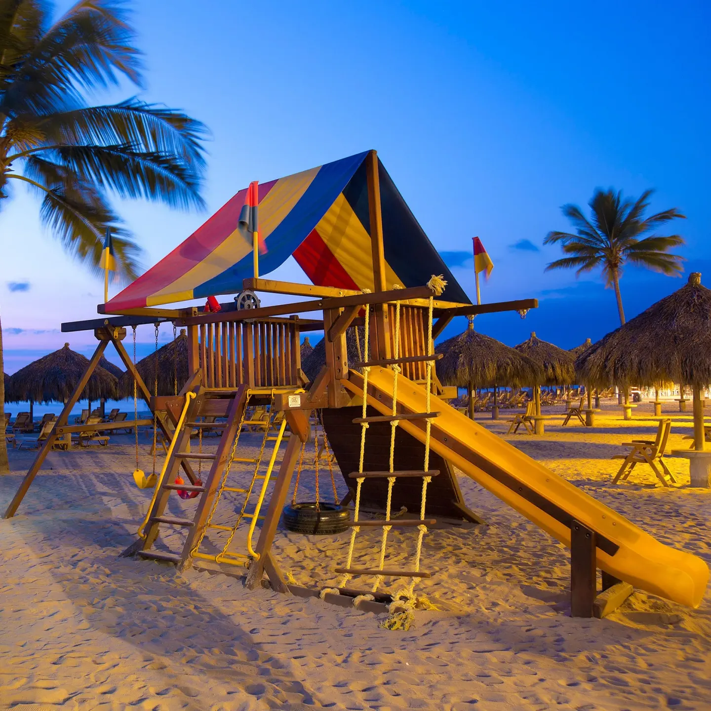 Beach playground at Paradise Village Beach Resort and Spa with slide, swings, palapas, and palms at dusk.