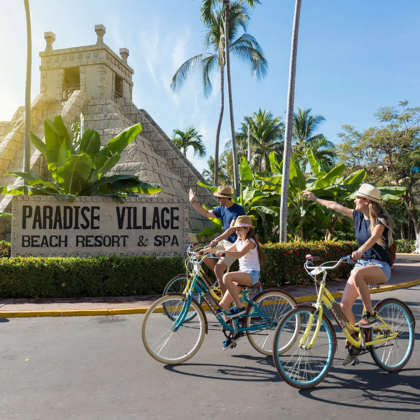 Family bicycling past the entrance sign at Paradise Village Beach Resort and Spa.