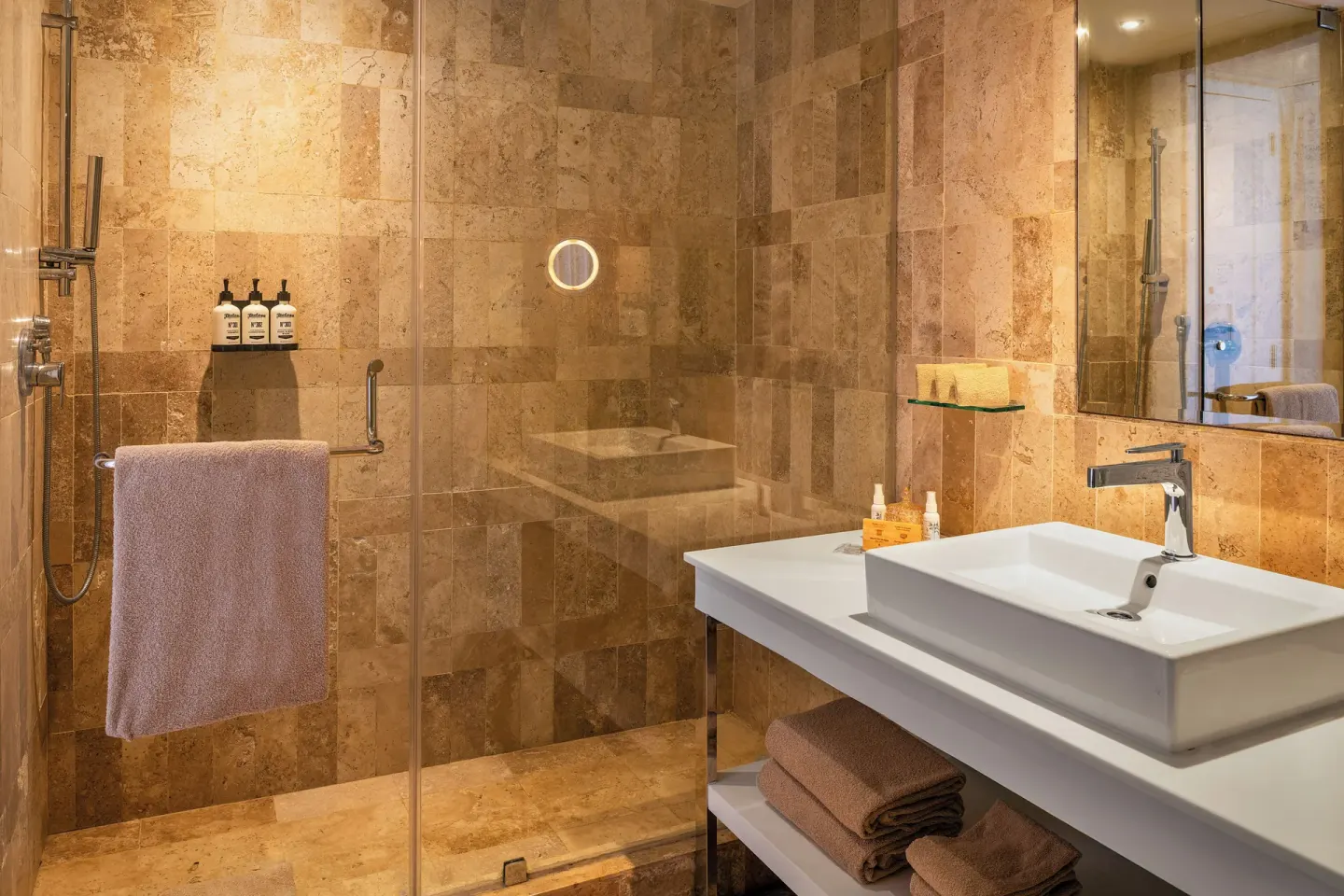 Glass-walled shower and white sink vanity in a warm stone bathroom at Paradisus Cancún.