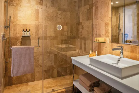Glass-walled shower and white sink vanity in a warm stone bathroom at Paradisus Cancún.
