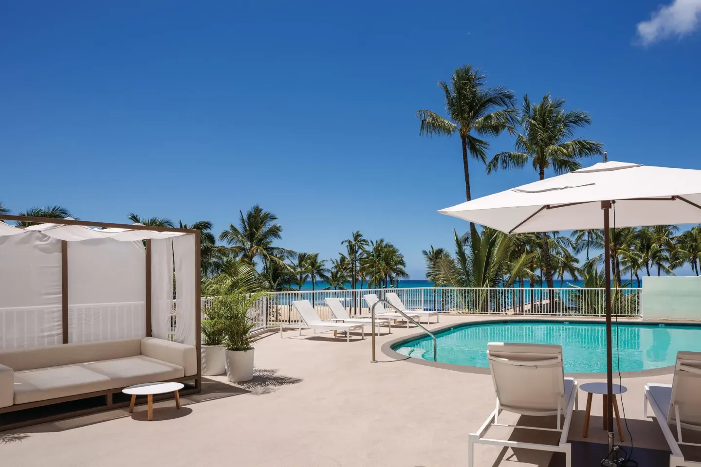 Pool deck with cabana seating, palm trees, and ocean beyond at Park Shore Waikiki.