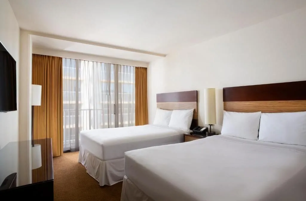 Standard Room with two white beds, bedside lamp, TV, and balcony curtains at Park Shore Waikiki.