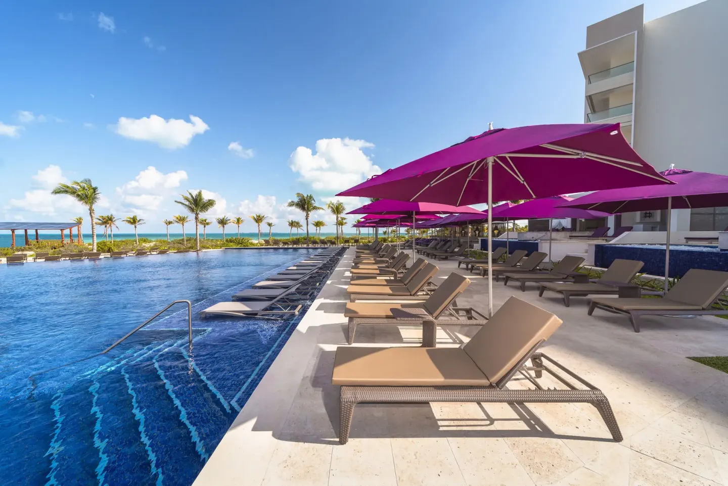 Poolside lounge chairs under purple umbrellas by the ocean at Planet Hollywood Cancun.