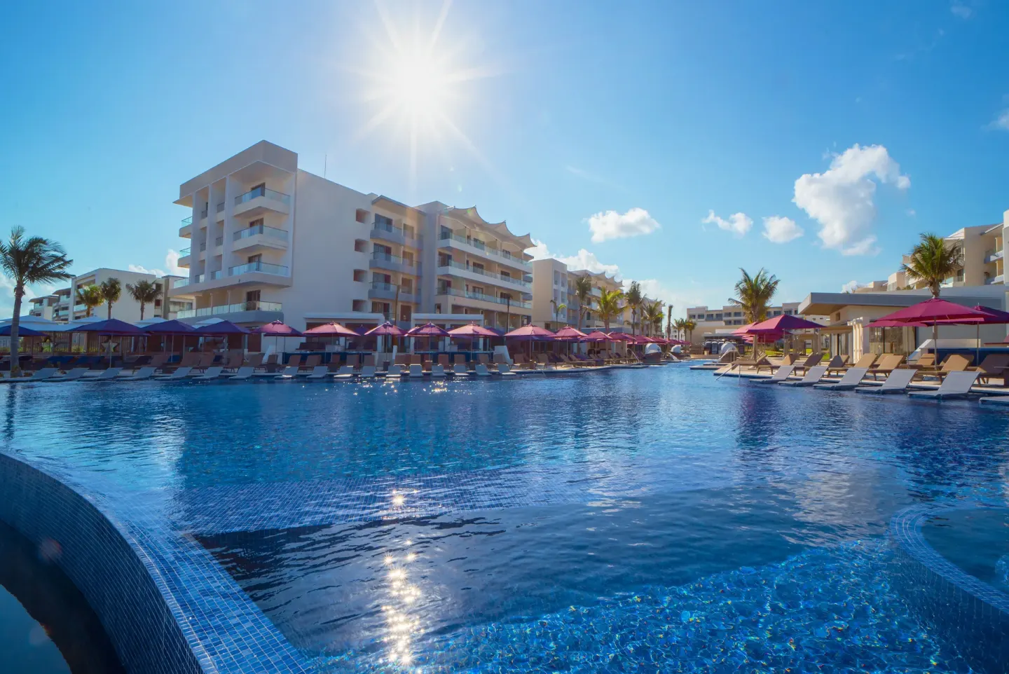 Sunlit main pool with resort buildings and rows of pink umbrellas at Planet Hollywood Cancun.