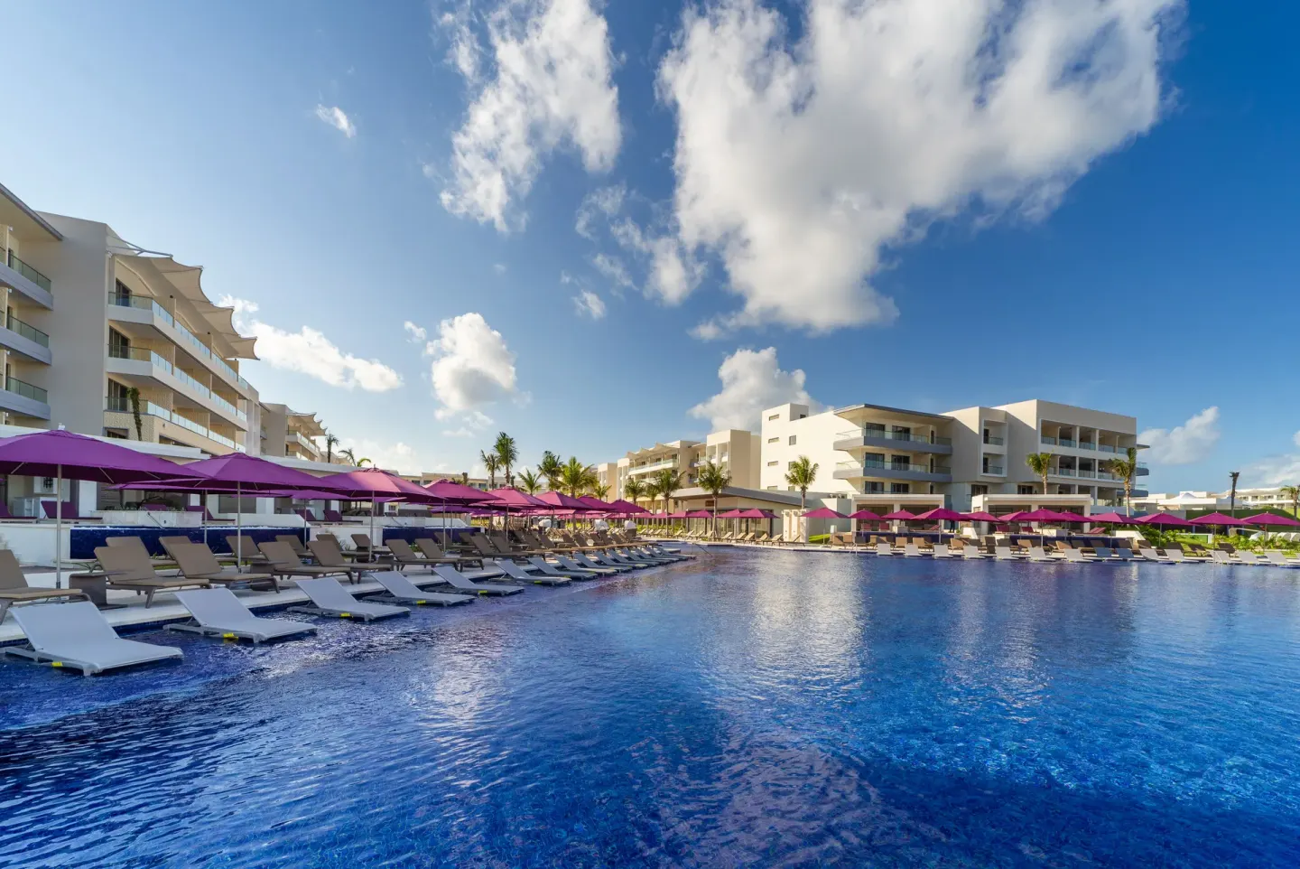 Wide resort pool bordered by loungers, pink umbrellas, and white buildings at Planet Hollywood Cancun.