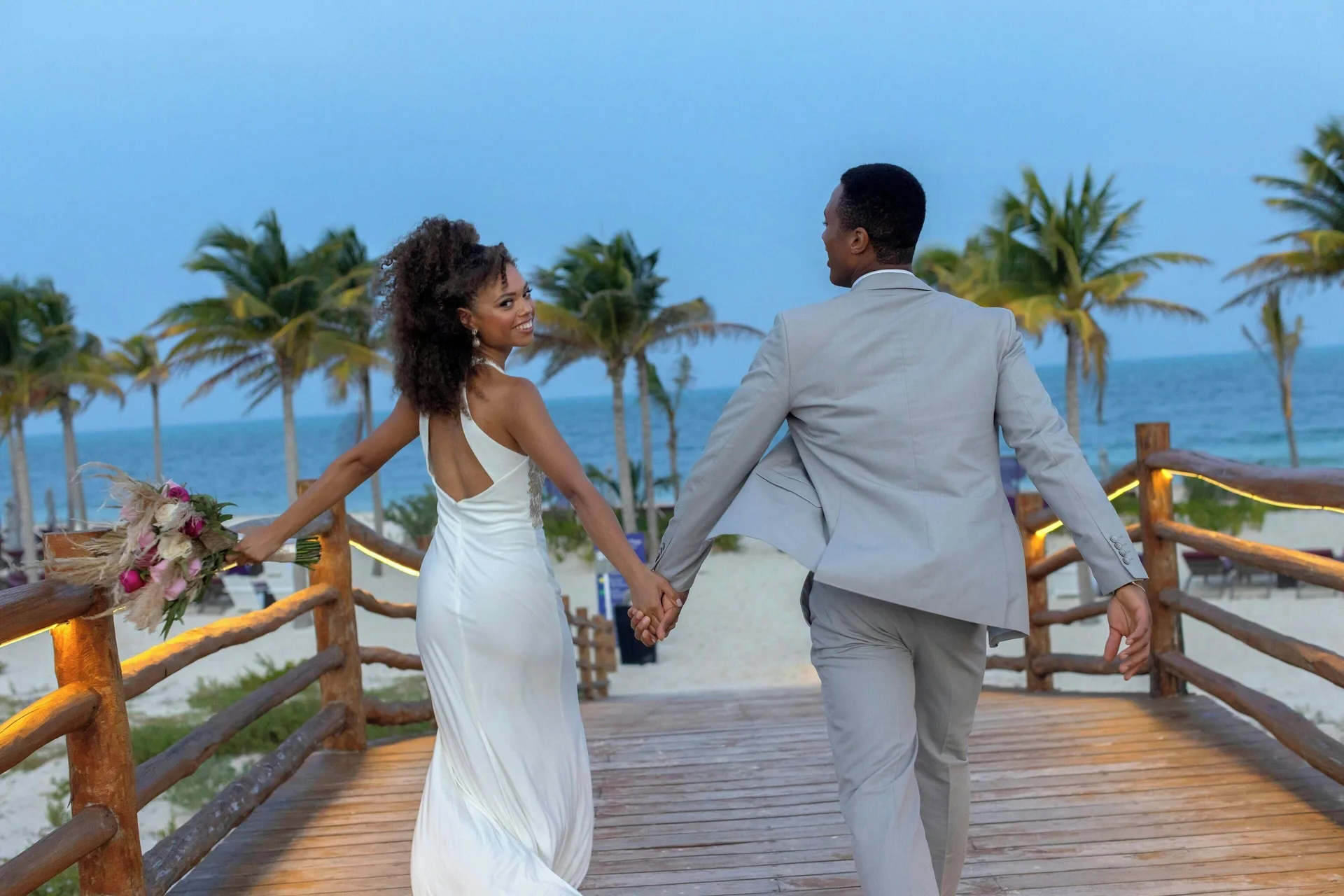Newlyweds walk hand in hand on a wooden beach walkway at Planet Hollywood Cancun.