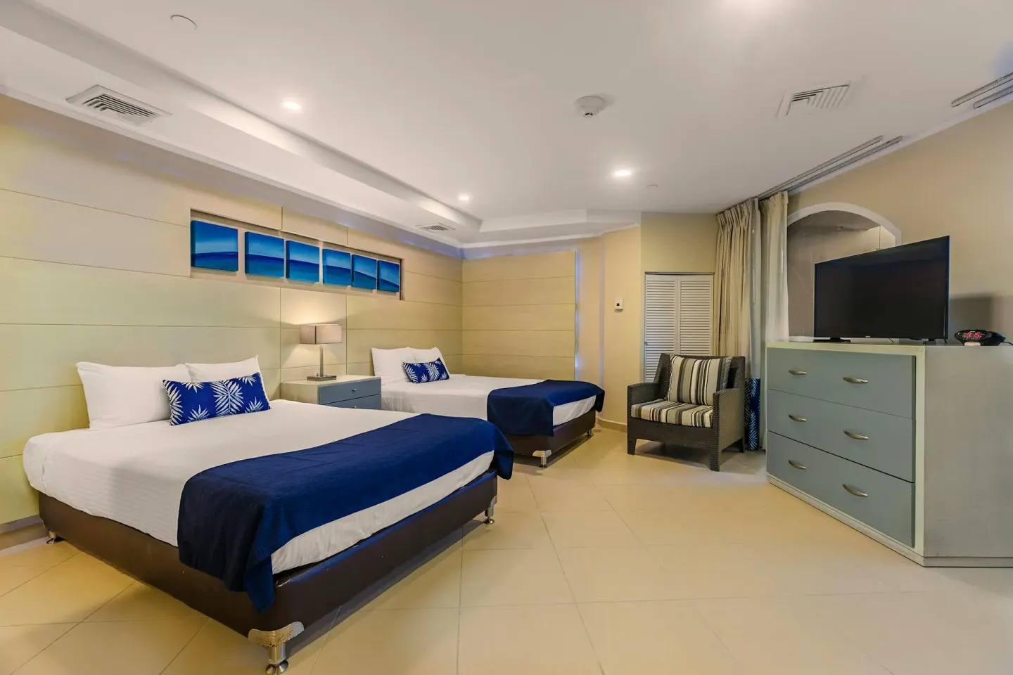 Spacious Spa Lanai Suite bedroom with two beds, ocean art panels and lounge chair at Playa Linda Beach Resort.