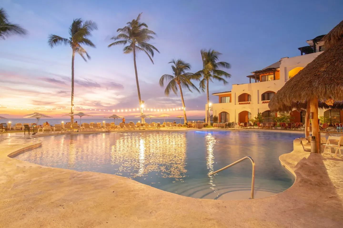 Evening pool at Plaza Pelicanos Club Beach Resort with palms, string lights, and guest room building.