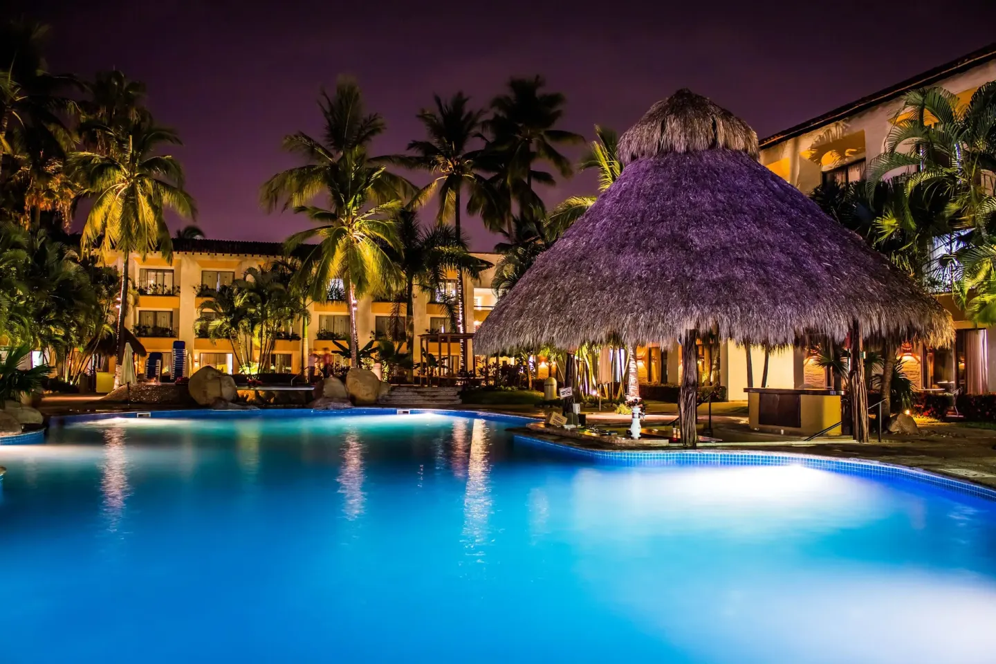 Illuminated pool and thatched palapa at night at Plaza Pelicanos Club Beach Resort.