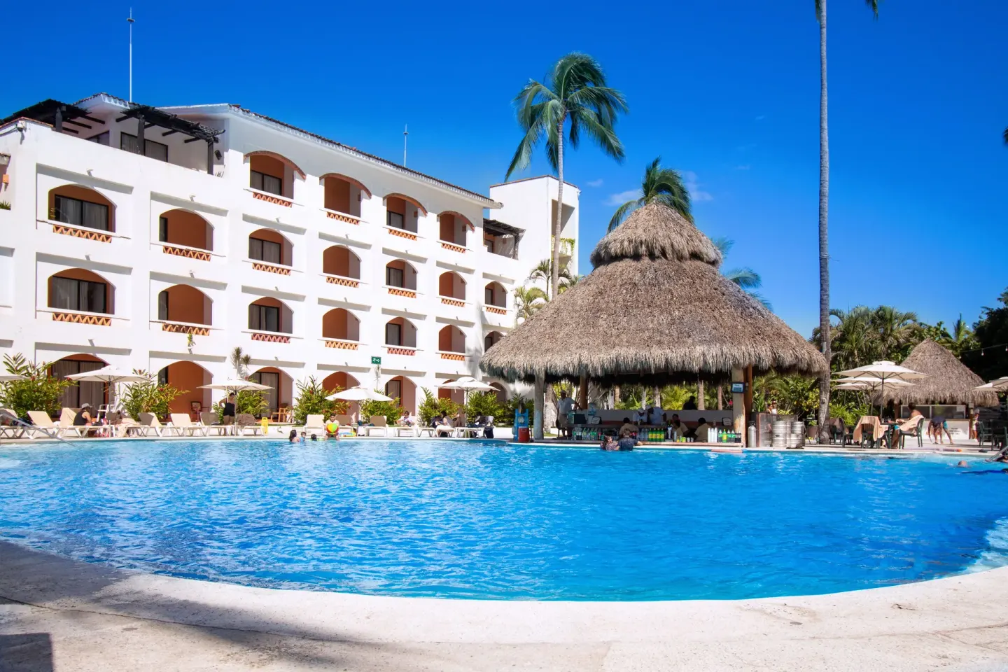 Large pool at Plaza Pelicanos Club Beach Resort with a thatched swim-up bar and guest room building.