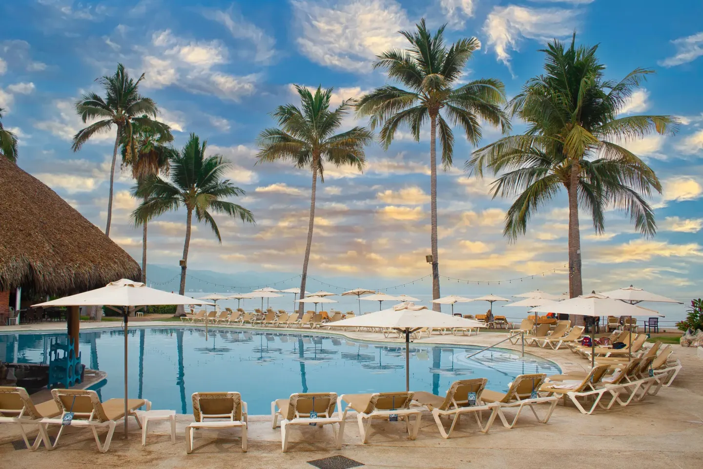 Resort pool at Plaza Pelicanos Club Beach Resort with palms, loungers, umbrellas, and a dramatic sky.