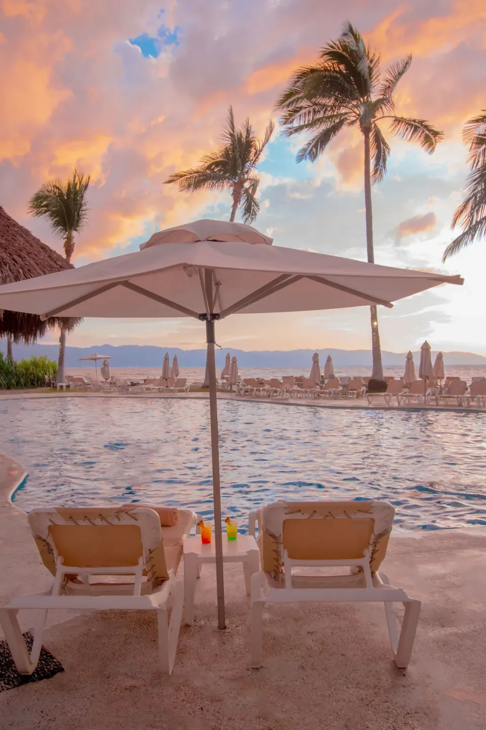 Poolside loungers under an umbrella at Plaza Pelicanos Club Beach Resort during sunset.