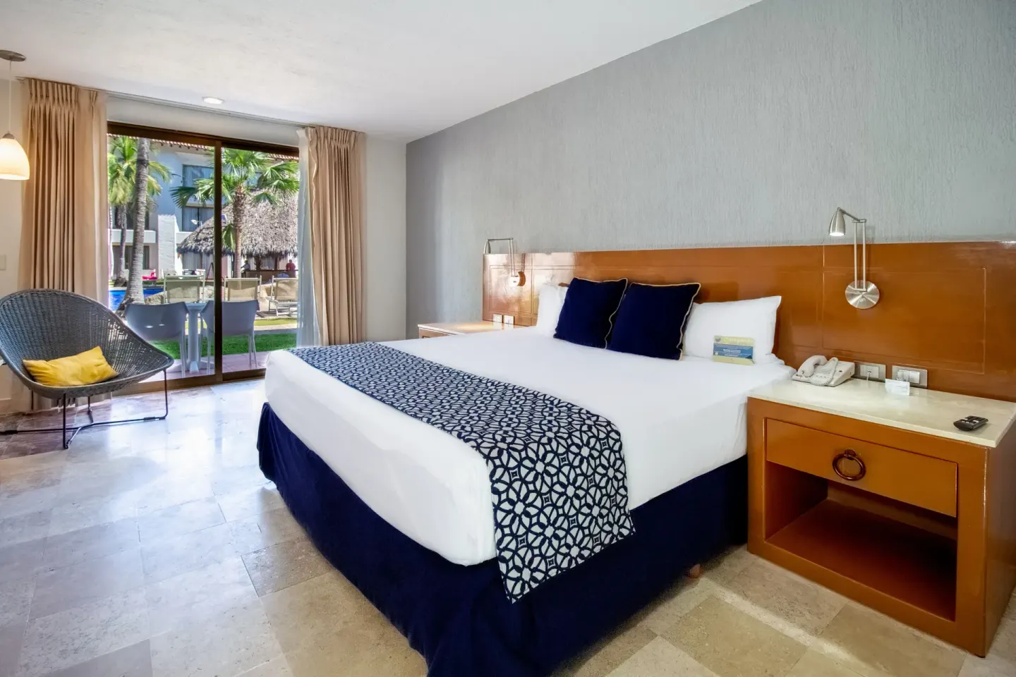 Superior Room king bed at Plaza Pelicanos Club Beach Resort beside nightstand and patio seating beyond.