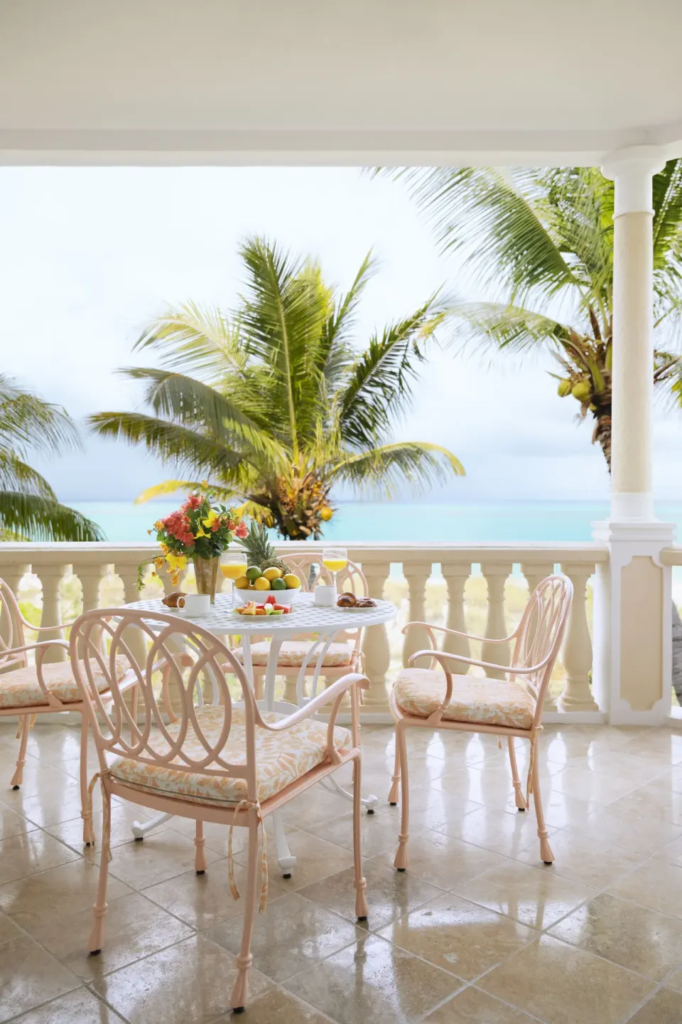 Covered balcony with pink dining chairs, fruit, flowers, palms, and sea view at Point Grace.
