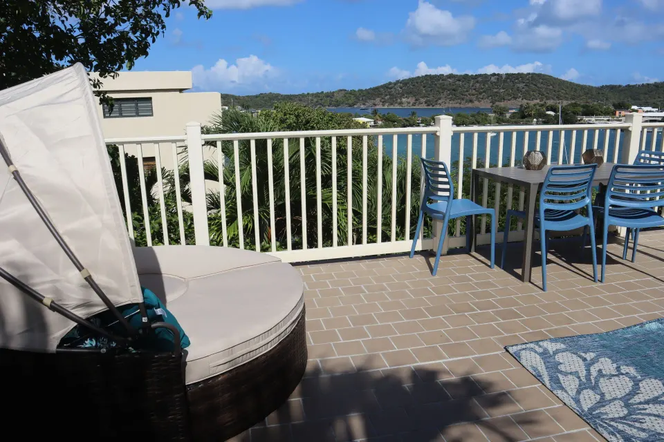 Studio terrace with canopy daybed, blue dining chairs, white railing, and water view at Point Pleasant Resort.