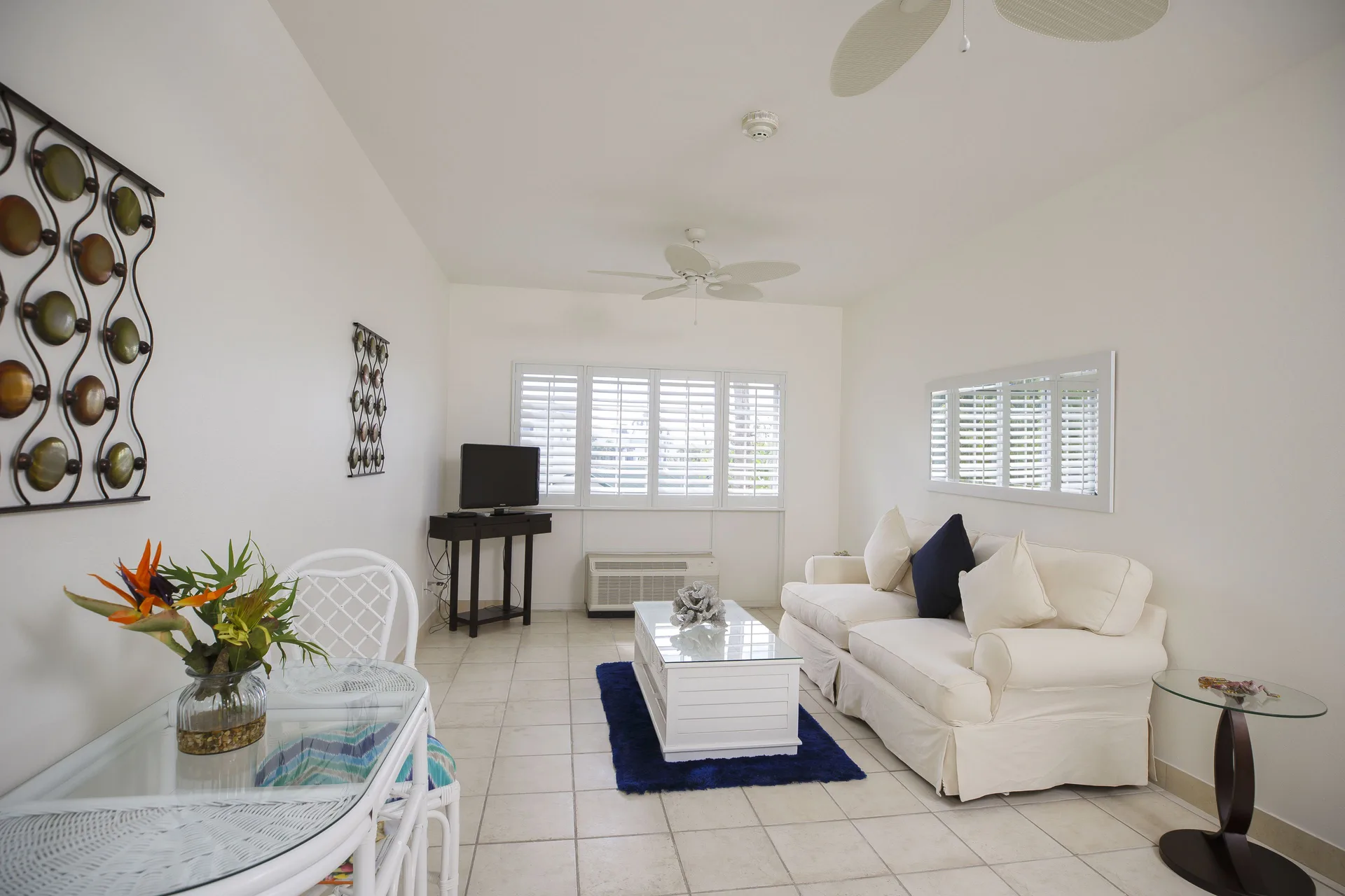 Living room with white sofa, TV, dining table, and shuttered windows at Ports of Call Resort.