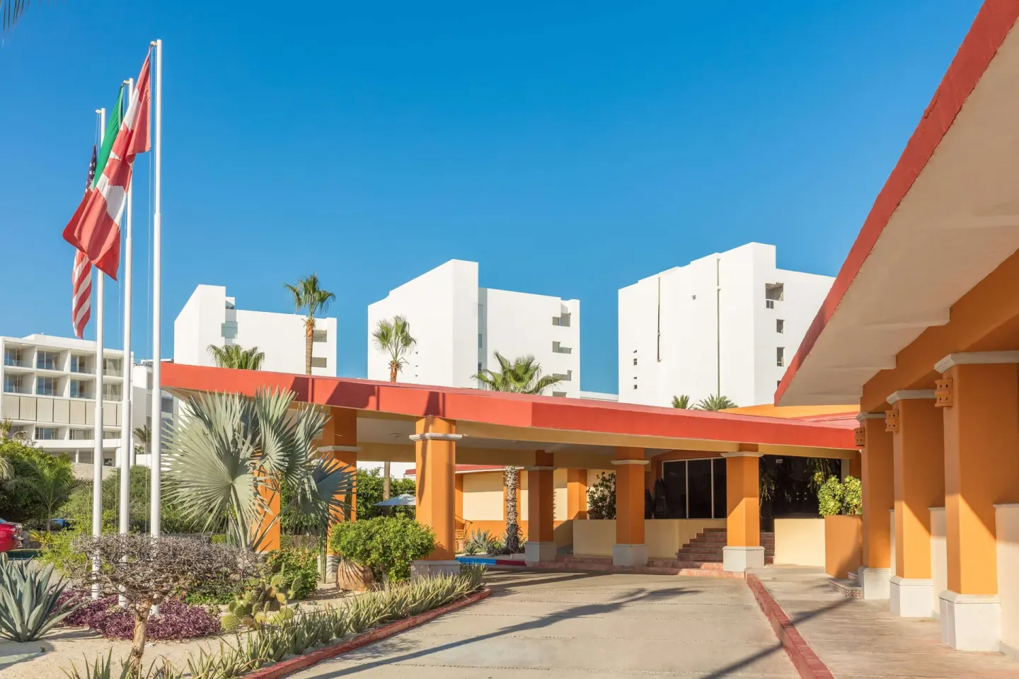 The entrance drive at Posada Real Los Cabos has orange columns, flags, palms, and white buildings.