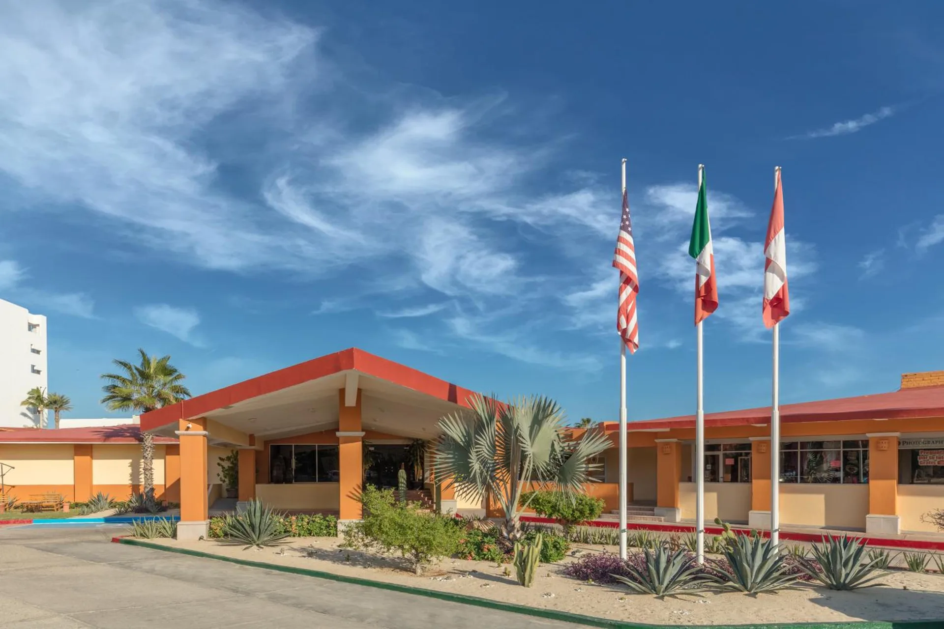 The front exterior of Posada Real Los Cabos shows flagpoles, orange buildings, palms, and desert plants.