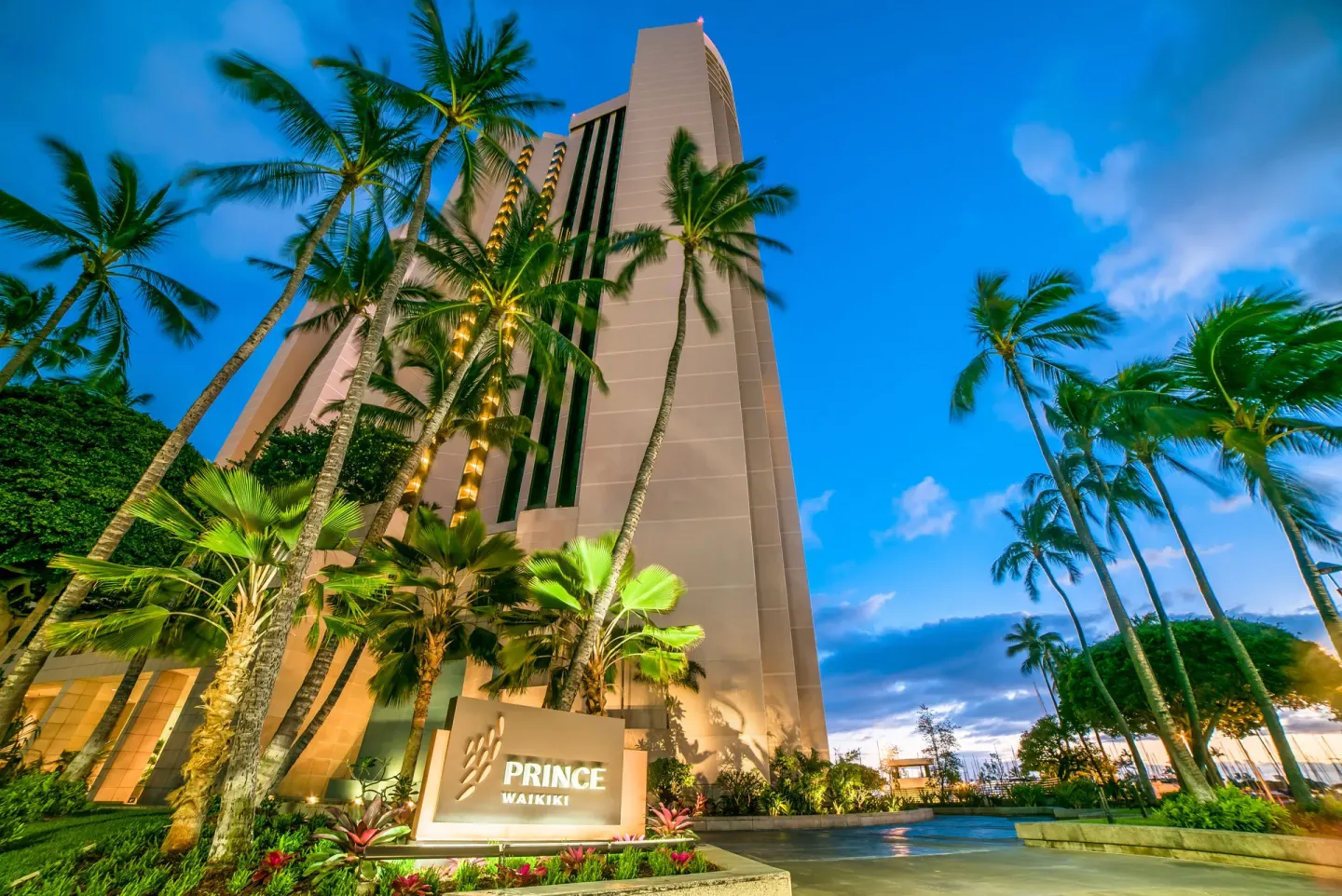 The Prince Waikiki tower rises above palm trees and a lit entrance sign at dusk.