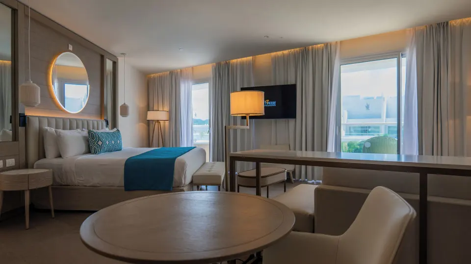 Wide view of the king bed, dining table, TV, and balcony windows at Princess Grand Jamaica Resort.
