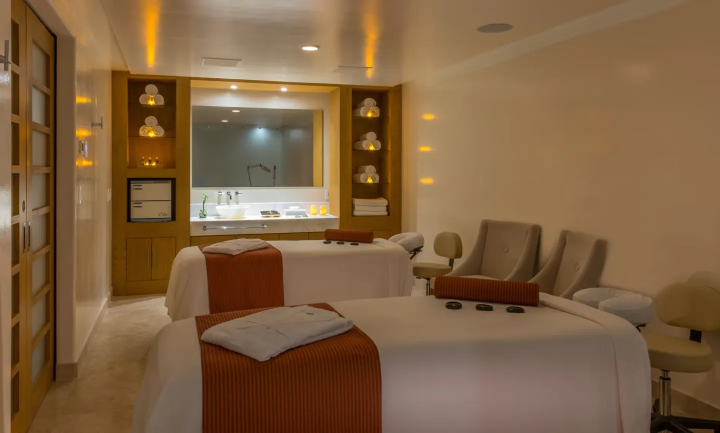 Spa treatment room with two massage tables, lounge chairs, shelves, and vanity at Pueblo Bonito Pacifica Golf & Spa Resort.