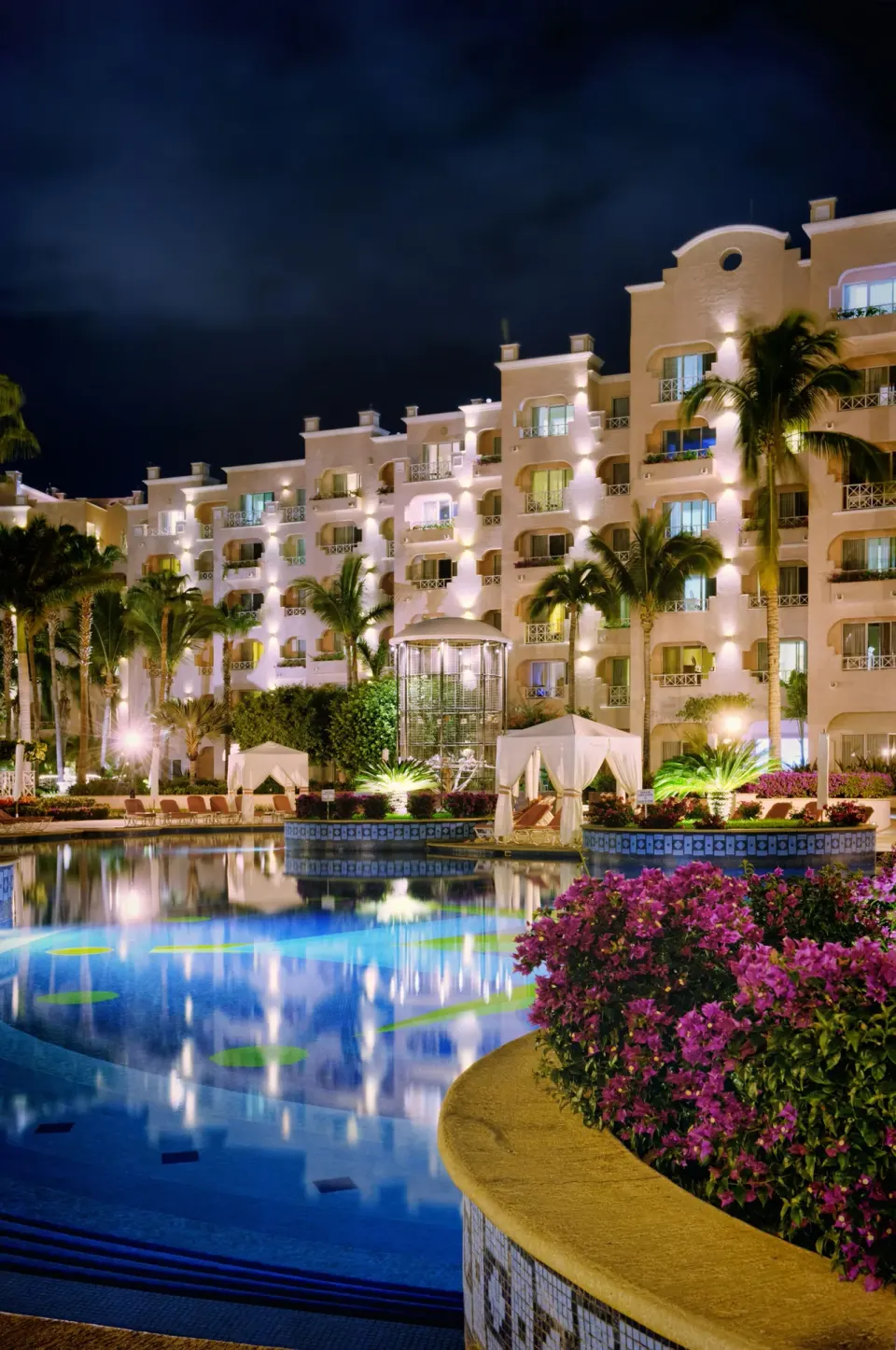 Nighttime pool, illuminated guest buildings, palms, and flowers at Pueblo Bonito Rose Resort and Spa.