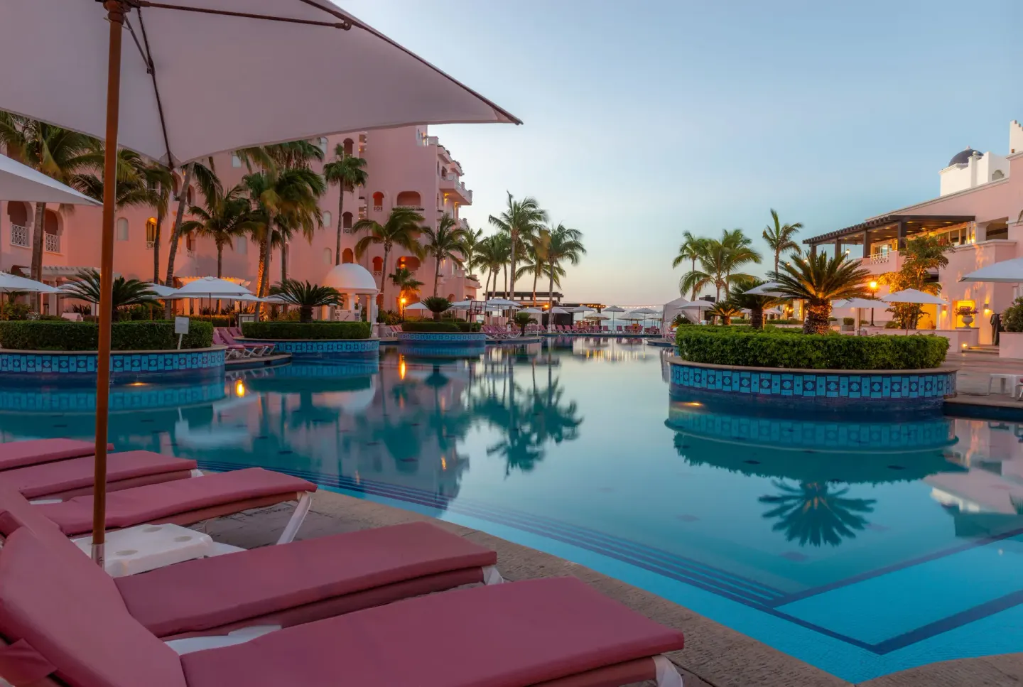 Poolside pink loungers, umbrellas, palms, and resort buildings at Pueblo Bonito Rose Resort and Spa.
