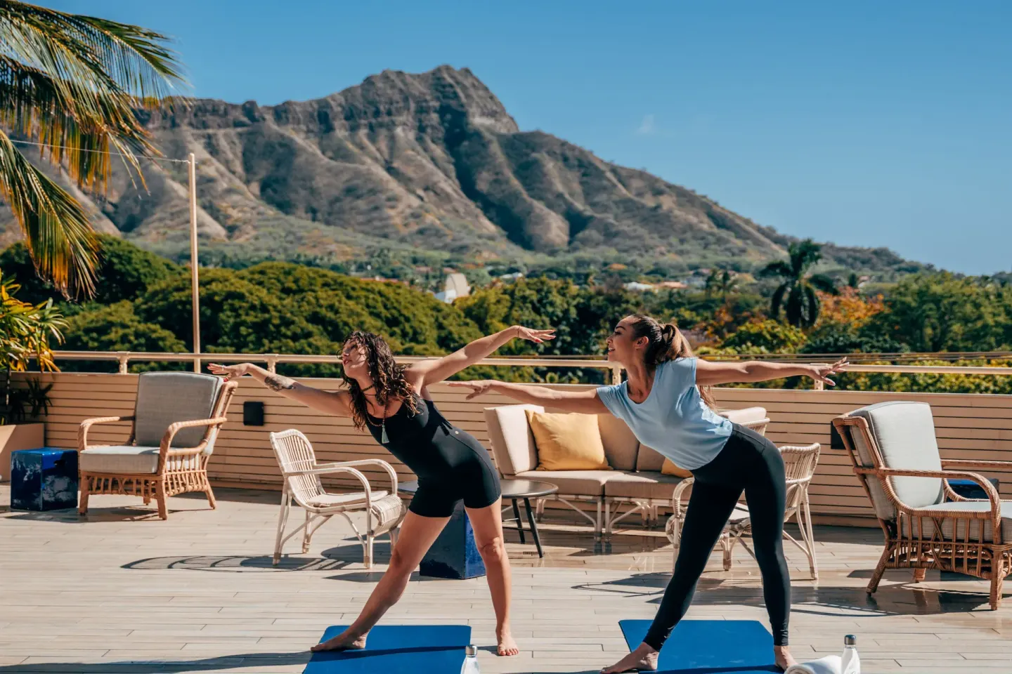 Two people practice yoga on an outdoor deck with Diamond Head beyond Queen Kapiolani Hotel.