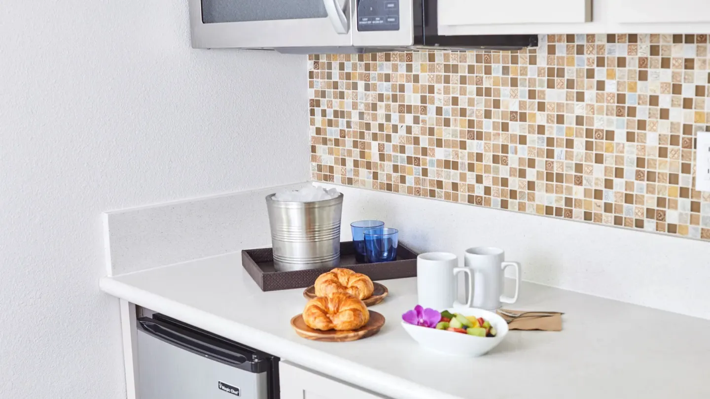 Junior Suite kitchenette counter with microwave, mini fridge, mugs, and snacks at Queen Kapiolani Hotel.