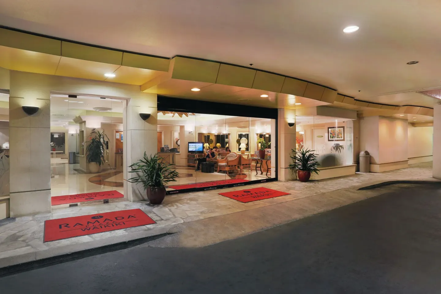 Covered entrance and lobby windows at Ramada Plaza Waikiki with red mats and seating inside.