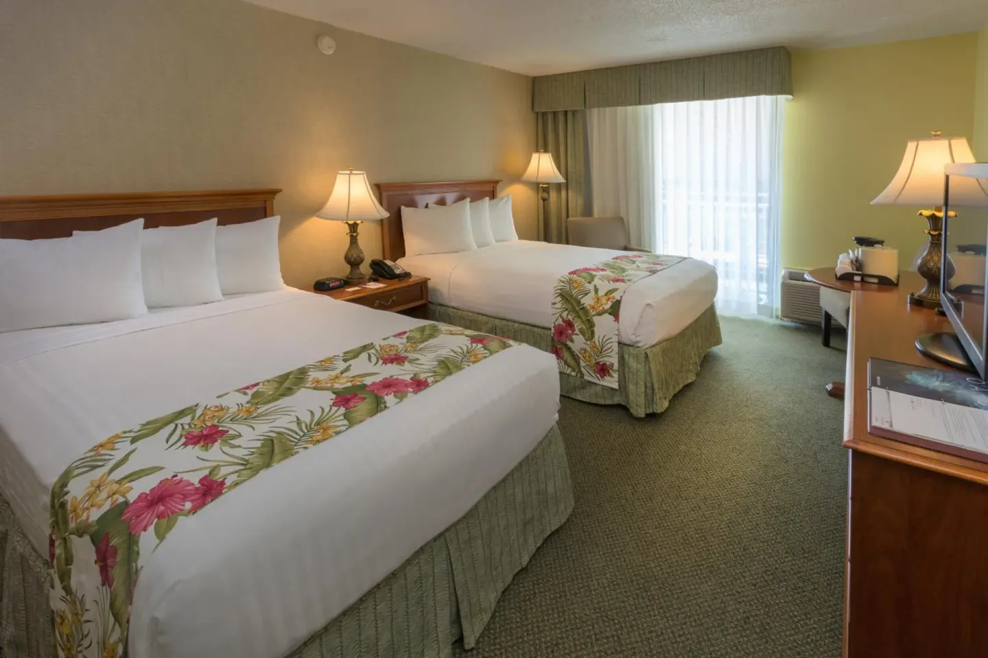 Two-bed guest room with desk, lamps, and curtained balcony door at Ramada Plaza Waikiki.