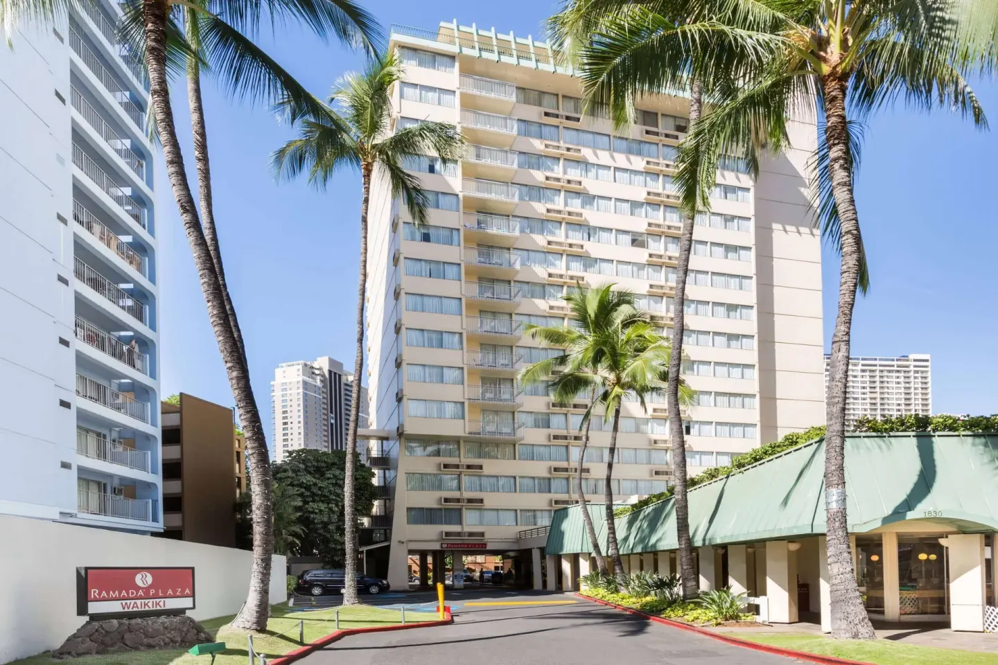 Exterior tower and driveway at Ramada Plaza Waikiki framed by palm trees and the hotel sign.