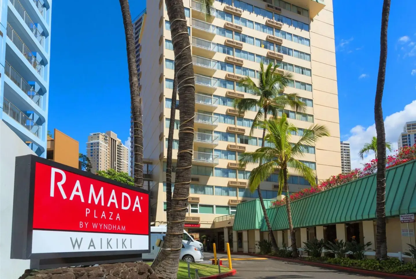 Close view of the Ramada Plaza Waikiki sign with palms, driveway, and hotel tower behind it.