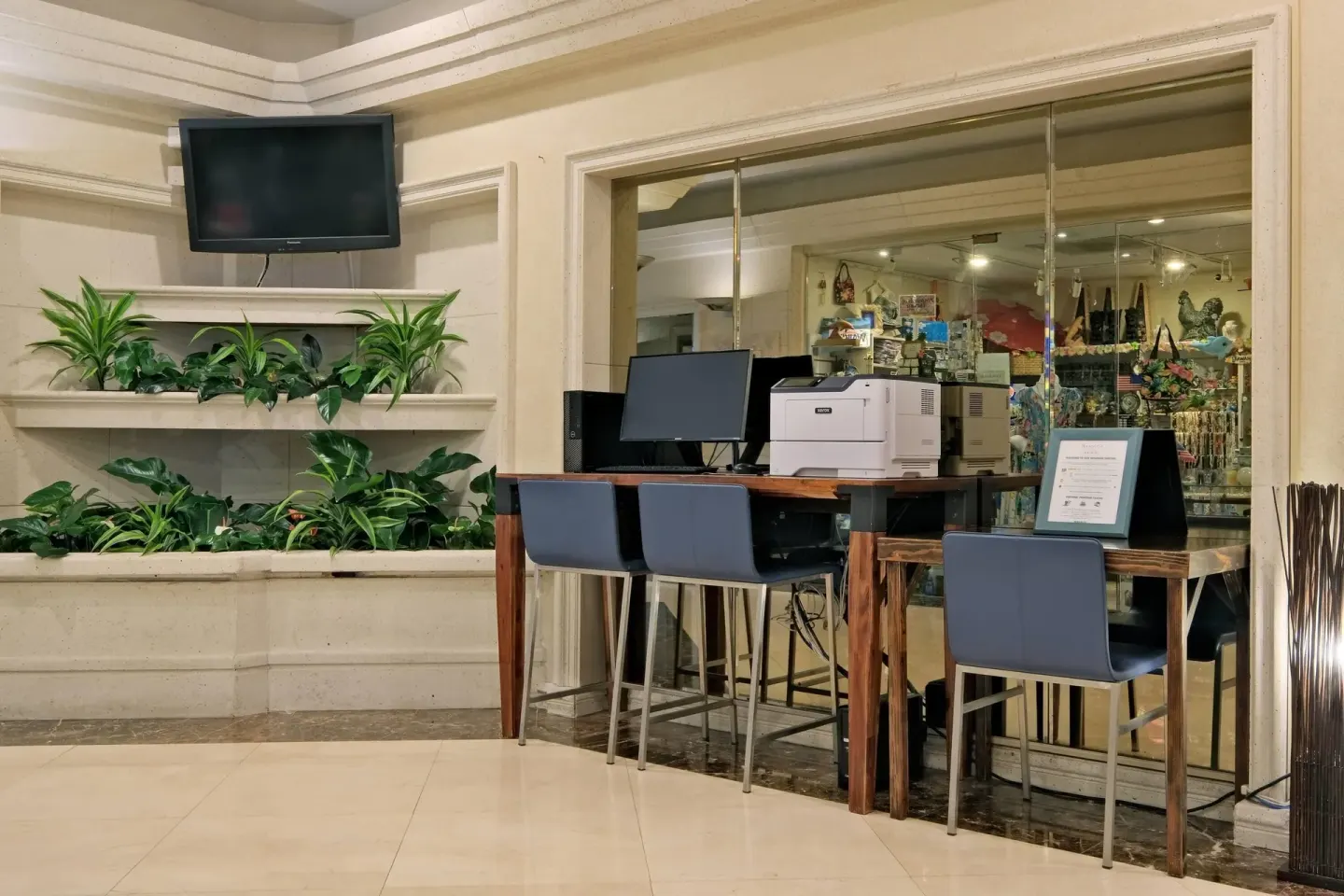 Lobby workstation with stools, printer, plants, and a wall TV at Ramada Plaza Waikiki.