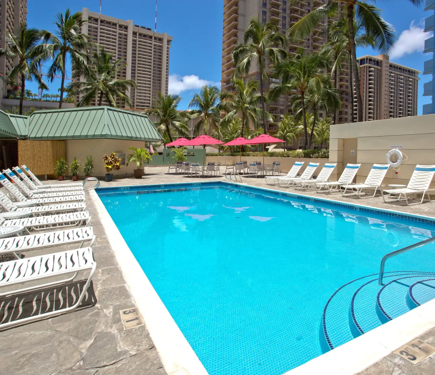 Outdoor pool at Ramada Plaza Waikiki with lounge chairs, red umbrellas, palms, and city towers.