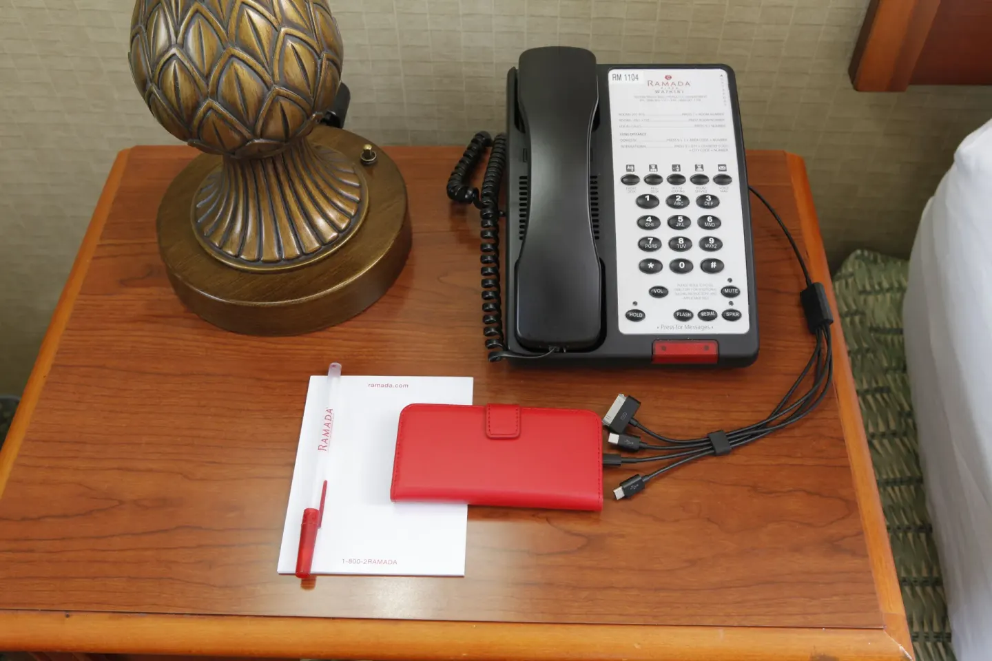Bedside table with phone, lamp, notepad and charging cables in a room at Ramada Plaza Waikiki.