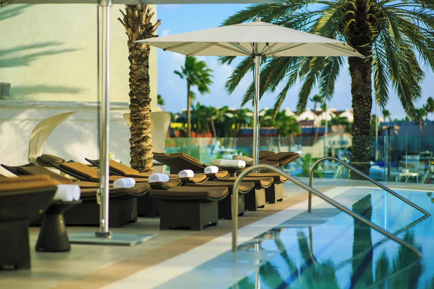 Poolside loungers under umbrellas and palms at Renaissance Wind Creek Aruba Resort.