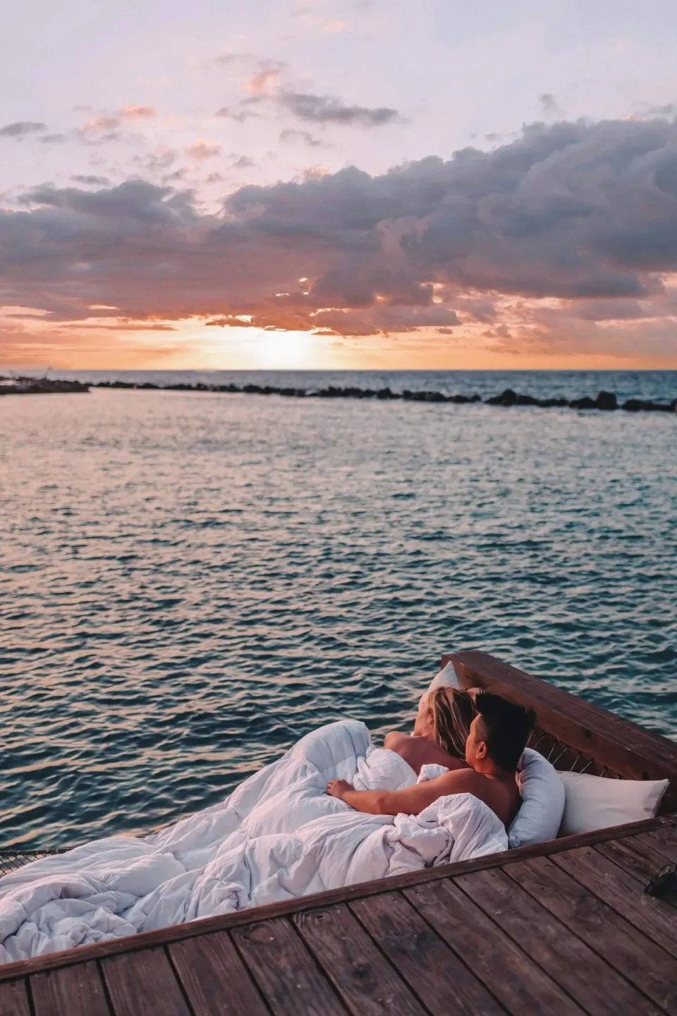 A couple rests in a waterfront hammock bed at sunset at Renaissance Wind Creek Aruba Resort.