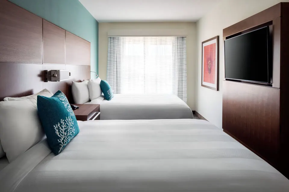 Two-bed suite bedroom at Residence Inn Maui Wailea with teal pillows, wood headboard, and TV wall.