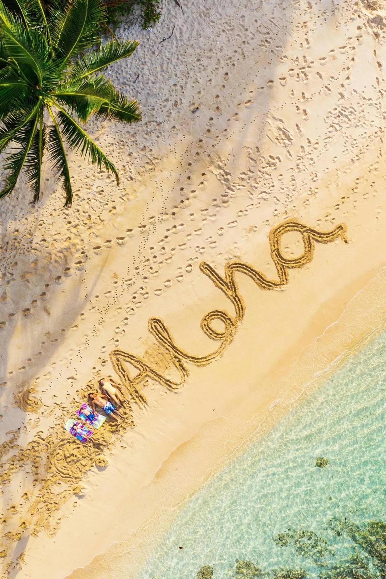 Aerial beach scene with Aloha written in sand near turquoise water.