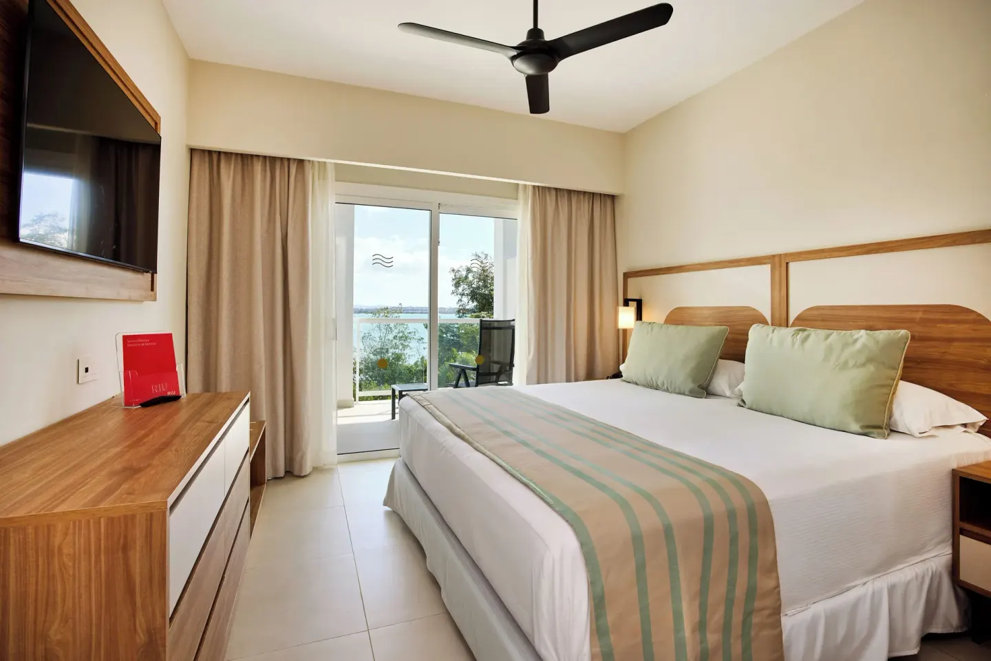 Ocean-view bedroom at Riu Negril with a large bed, balcony doors, TV, and dresser.