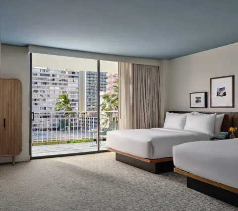 Deluxe Room with two double beds, balcony doors, and city buildings at Romer House Waikiki.