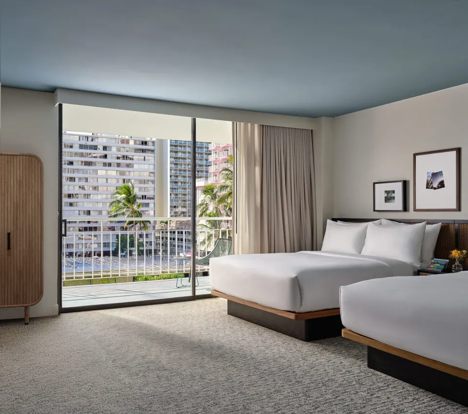 Deluxe Room with two double beds, balcony doors, and city buildings at Romer House Waikiki.