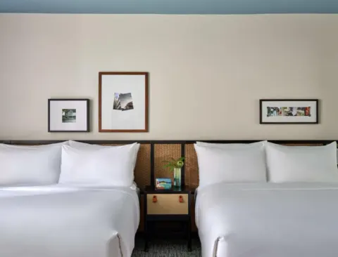 Deluxe Two Queens ADA room at Romer House Waikiki with two white queen beds and framed wall art.