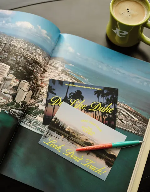 Standard room tabletop at Romer House Waikiki with open photo book, postcards, pen, and coffee mug.
