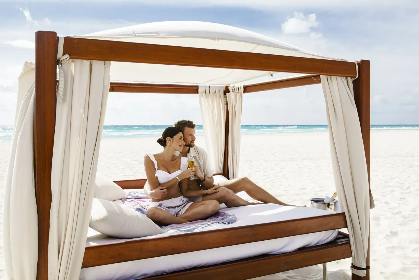 Couple relaxes with drinks on a curtained beach daybed at Royal Hideaway Playacar.