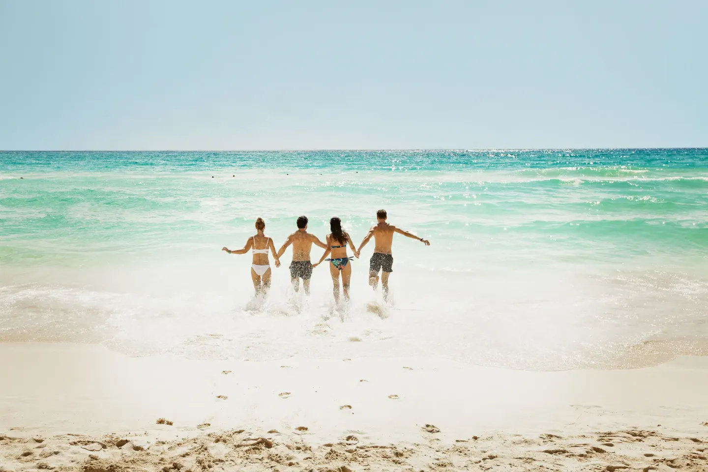 Four people run into turquoise surf from the beach at Royal Hideaway Playacar.
