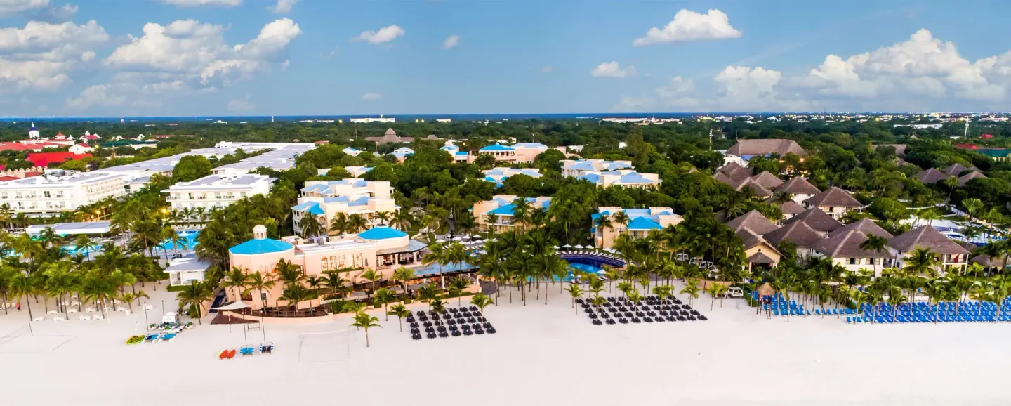 Wide aerial view of Royal Hideaway Playacar with beachfront loungers, pools, and blue-roofed buildings.