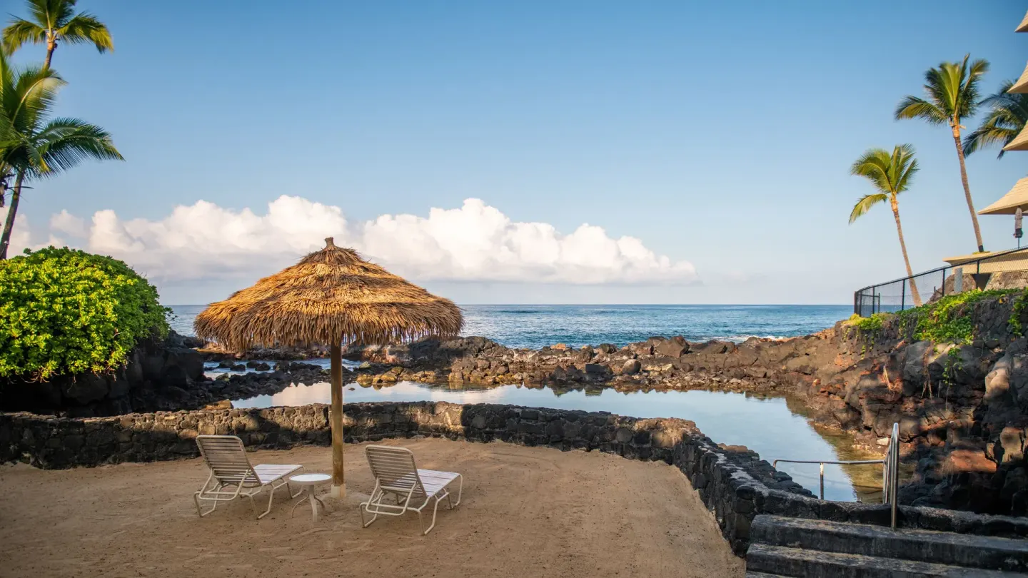 Two lounge chairs sit under a thatched umbrella by the lagoon at Royal Kona Resort.