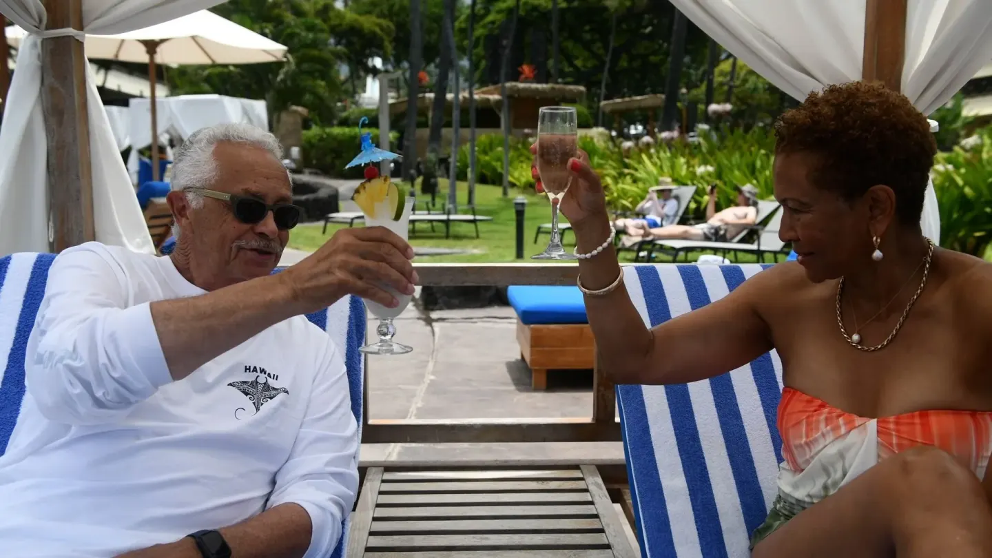Guests toast drinks on striped lounge chairs by the pool at Royal Kona Resort.